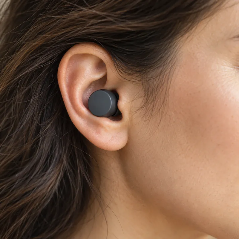 Close view of an ear wearing a graphite earbud. Marks show the earbud's speaker, a heartbeat at the entrance of the sealed ear canal and a tap on the earbud.